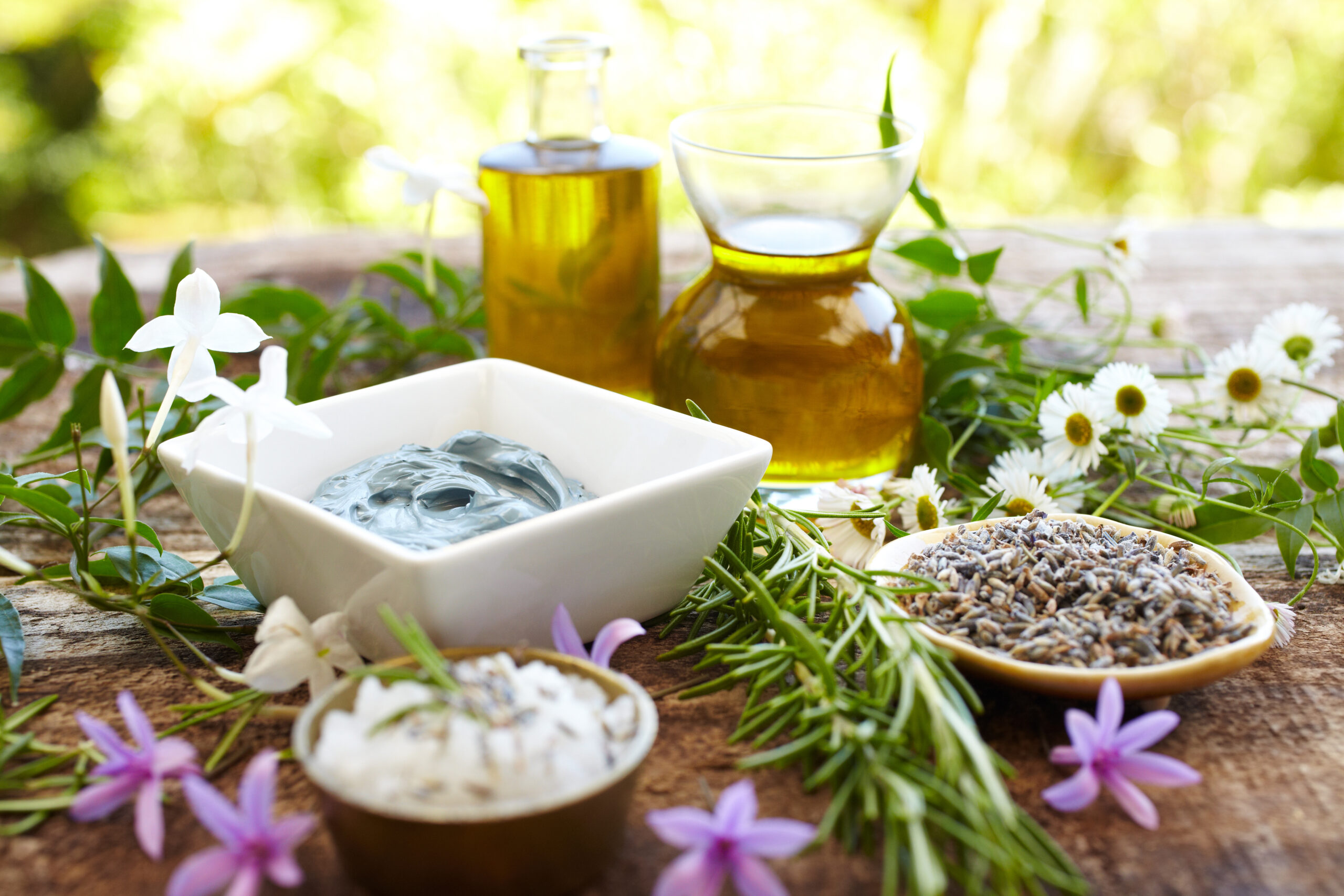 Spa still life of herbs, oil, blue chamomile body mud mask, rosemary, salt scrub exfoliant, fresh wild flowers on a distressed wood surface shot from above.  Square shot.